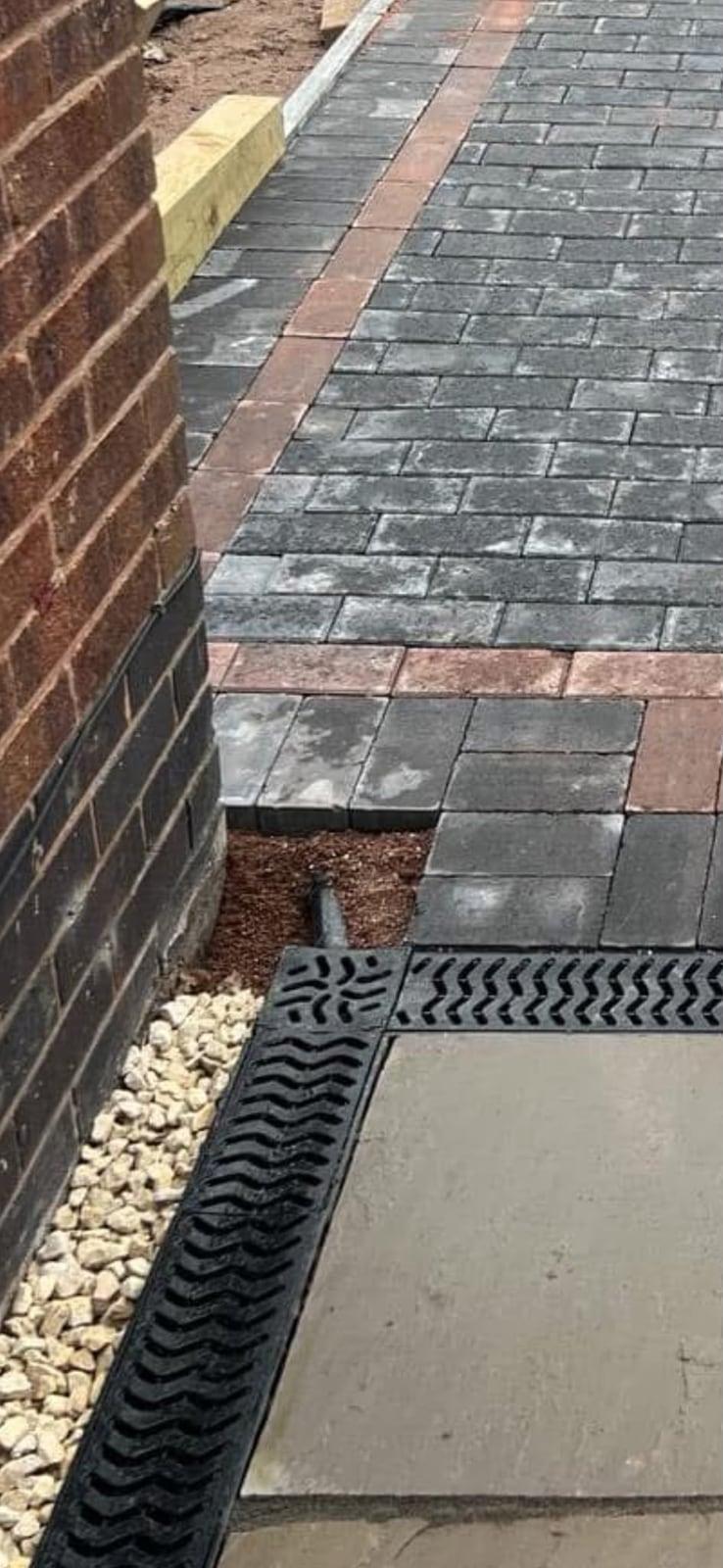 Drainage channel running along new block paving beside the house wall, with a gravel strip