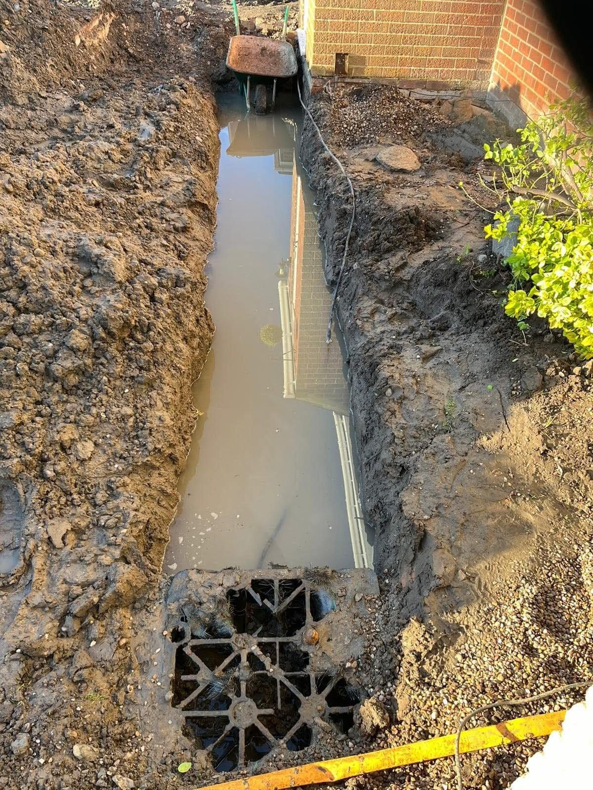 Trench dug alongside a house, holding rainwater, with soakaway crates going in at the end